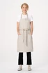 A light beige bib apron with a tie waist and front pockets, worn by a model against a plain background.