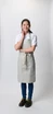 A woman wearing a light grey Lockharte bib apron, paired with a white shirt and dark blue jeans, smiles confidently.
