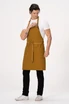 A male model wears a mustard yellow Lockharte Bib Apron over a white t-shirt and black pants, featuring a logo.