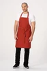 A bib apron in rust colour, featuring a waist tie, modelled by a man wearing a white t-shirt and black pants.