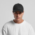 A black trucker cap with a structured front panel and mesh back, worn by a model in a white t-shirt.