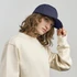 A woman wearing a navy cap and a cream-coloured sweatshirt, posing with one hand on her cap.