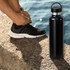A drink bottle in matte black stainless steel with a handle, placed on a stone surface near water.