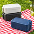 A stack of lunchboxes in navy, black, and white, sitting on a red and white checkered picnic blanket.