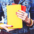 A person holds a yellow notebook along with a red notebook, wearing a denim jacket and colourful bracelets.