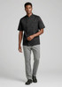 A model wearing a black short-sleeve chef jacket and grey pants, walking in a neutral background. The outfit features a logo.