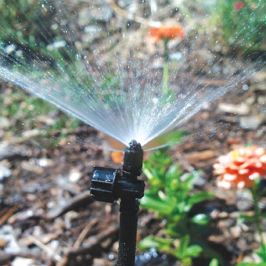 An in-use photo of the 90° Micro-Jet Sprayer on a stake, producing a wide fan spray over a flower bed.