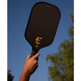 Hand holding the Angled Electrum Model E 13mm Pickleball Paddle with black paddle background and Electrum logo in gold into the air against blue sky and green trees.