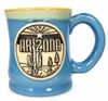 Blue glazed ceramic mug with a glossy finish, cream and yellow-green rim, rounded body, and curved handle. The front features a raised circular Southwestern emblem with “ARIZONA” lettering, a rising sun, a star, saguaro cactus, and desert vegetation.