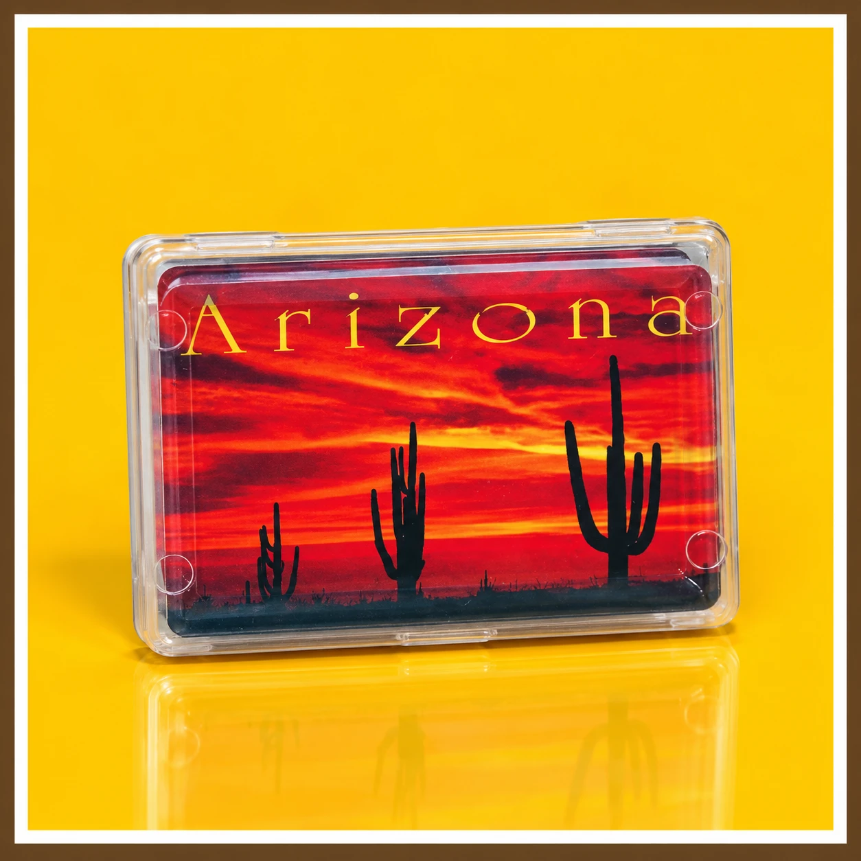 Arizona Sunset Playing Cards in a clear plastic case featuring a dramatic red-orange sunset, black saguaro cactus silhouettes, and golden Arizona lettering against a Southwestern desert sky.