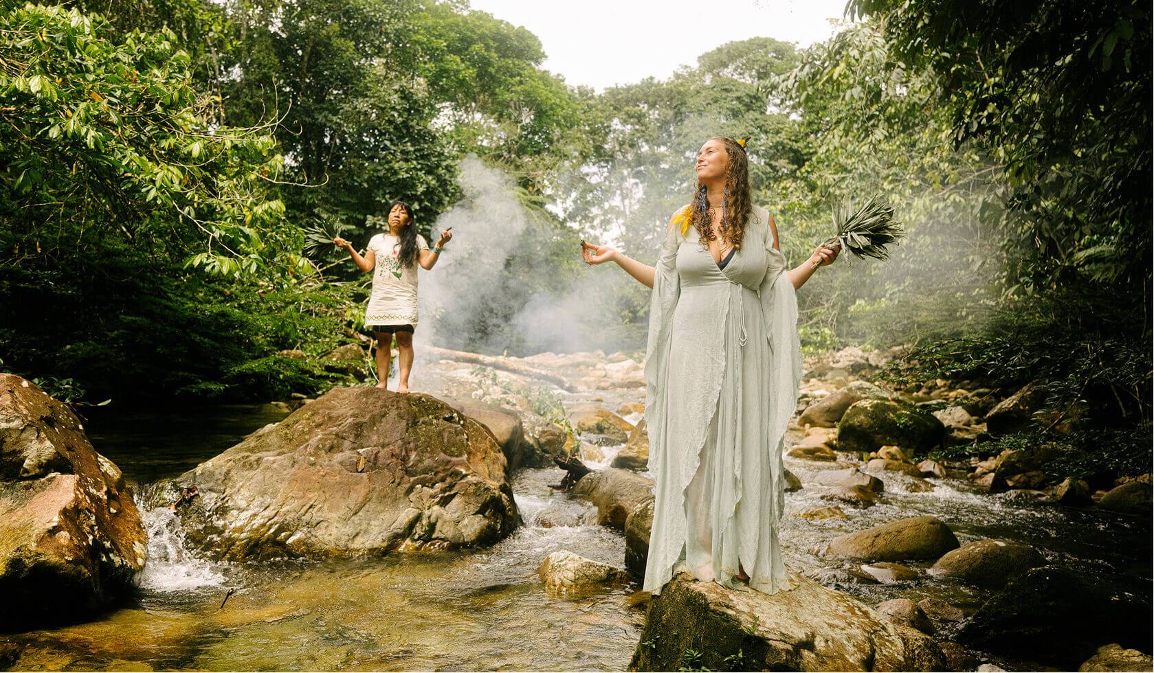 Two women standing on rocks by a forest stream surrounded by lush green tropical foliage, performing a sacred plant ceremony with smoke rising into the canopy