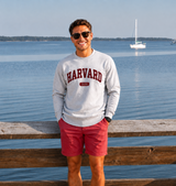 Harvard Nantucket fleece crewneck sweatshirt in heather grey with embroidered maroon Harvard 1636 lettering, shown in a coastal New England setting overlooking the water.