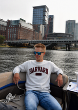 Harvard Nantucket fleece crewneck sweatshirt in heather grey with embroidered maroon Harvard 1636 lettering, shown in a coastal New England setting overlooking the water.