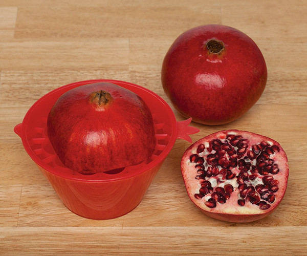 Half a pomegranate placed cut side down on the deseeder's grid over the bowl, on a wooden board beside a whole pomegranate and a cut half.