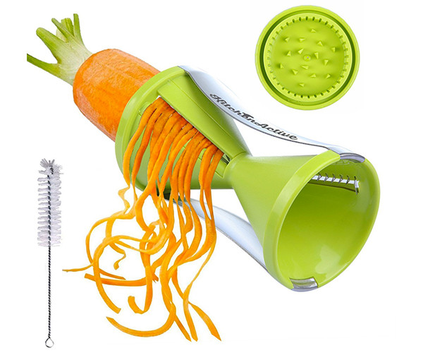 Handheld spiral slicer cutting a carrot into long noodle-like strands, shown with its gripper cap and a cleaning brush.