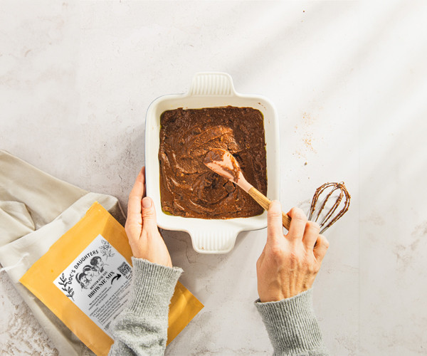 Hands spreading Better For You brownie batter in a white baking dish, with a whisk and the mix pouch beside it.