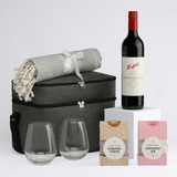 A drink bottle of red wine with two clear glasses, a grey blanket, and two packets of sweets in a stylish bag.