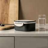 A black and white plastic lunch box on a kitchen countertop, beside a glass of water and a wooden cutting board.