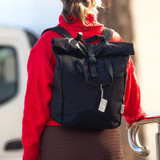 A black recycled canvas backpack tote worn on a person’s back, featuring a roll-top design and a fastening strap.