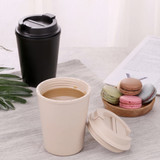 A reusable coffee cup in beige with a lid, next to a black cup and a plate of colourful macarons.