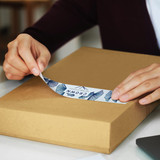 A person applying a blue and white sticker to a brown packaging box. The box is on a flat surface. A person applying a blue and white sticker to a brown packaging box. The box is on a flat surface.