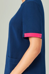 Unisex navy scrub top with pink accents on the sleeves, featuring a side pocket and a circular logo detail.
