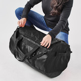 A black duffle bag is being held by a person in casual attire, positioned on a white background.