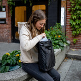 A woman interacting with a black Meridan Tote Backpack while sitting on a stone ledge with greenery around.