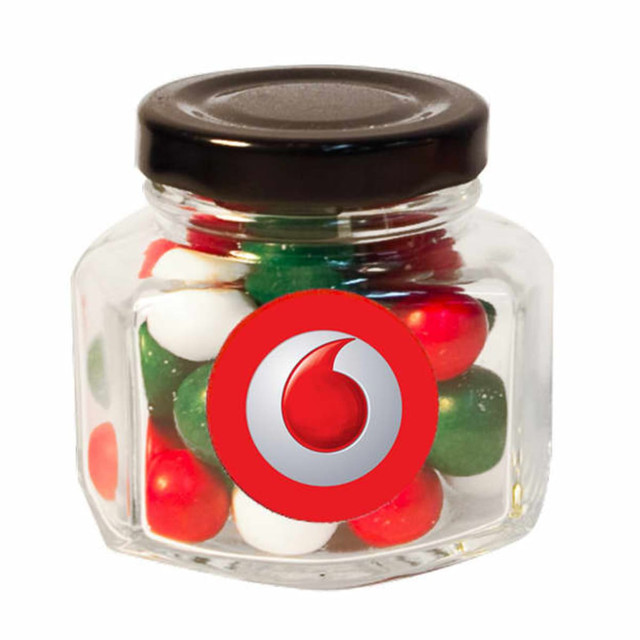 A clear glass jar with a black screw top filled with red, green, and white chocolate balls, featuring a logo.