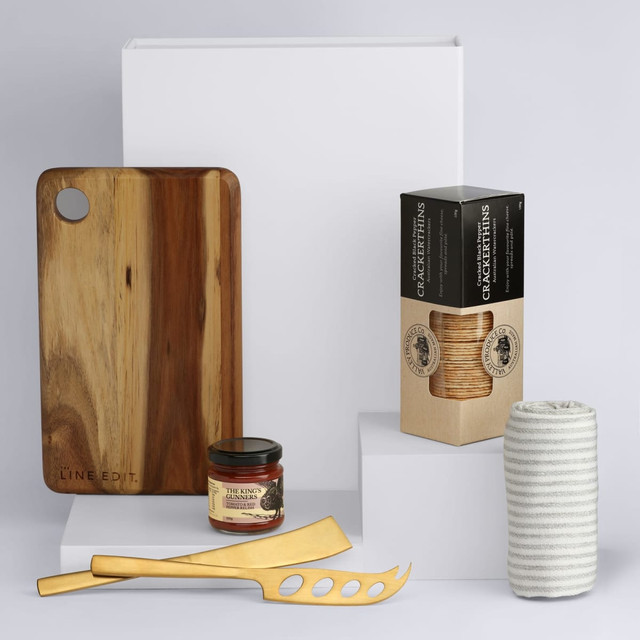 A wooden cheese board, slotted knife, spreader, jar of dip, and a box of crackers on display against a light background.