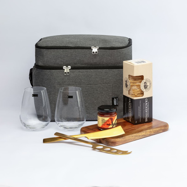 A grey carrying bag with two glasses, a jar, crackers, a cheese knife, and a wooden platter.