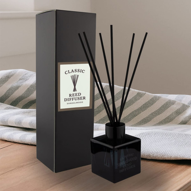 A black square reed diffuser with black reeds, packaged in a matching black box, sits on a wooden surface.