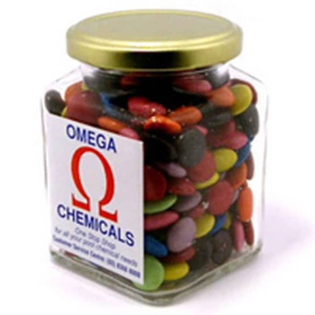A glass square jar filled with colourful choc beans, featuring a gold lid and a branded sticker on the front.