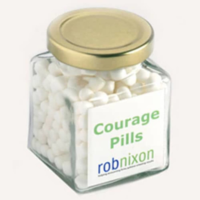 A square glass jar filled with white mints, features a gold lid and a branded sticker.