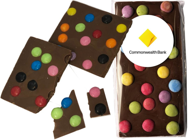 A milk chocolate bar with colourful round candies on top, partially broken, presented in a clear wrapper with a logo.