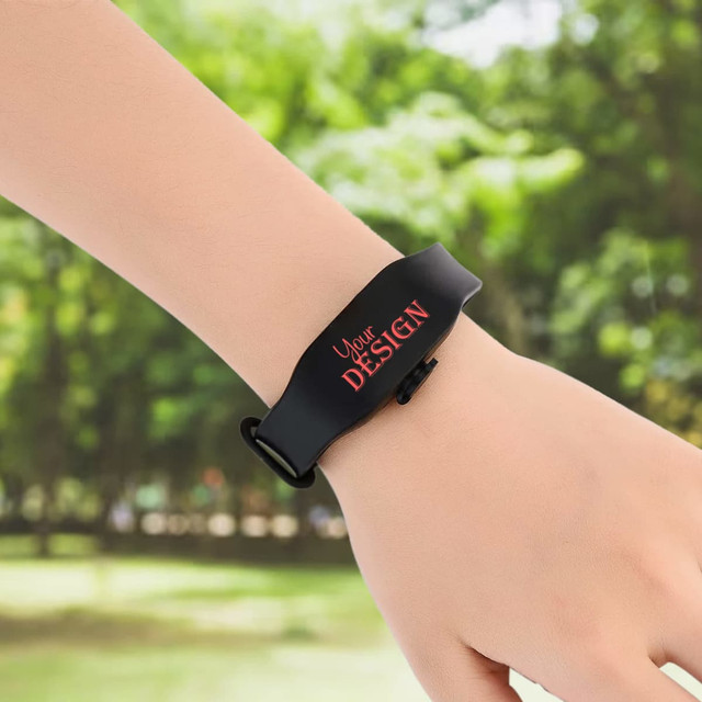 A black wristband hand sanitiser dispenser with a logo, worn on a person's wrist in a green outdoor setting.