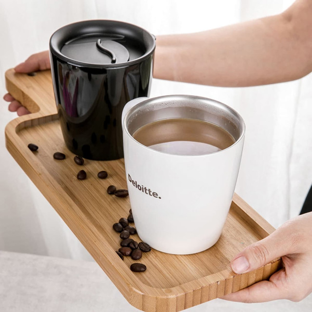A drinkware set featuring a black and a white coffee mug on a wooden tray, surrounded by coffee beans. Each mug has a lid and one features a logo.