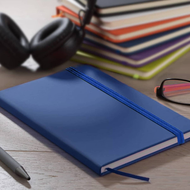 A blue A5 PU notebook with an elastic band, placed alongside headphones and glasses, with stacked notebooks in the background.
