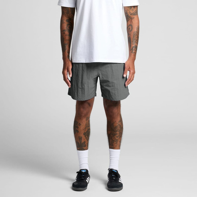 A person stands wearing grey swim shorts, paired with a white t-shirt and black sneakers. The shorts feature a logo.