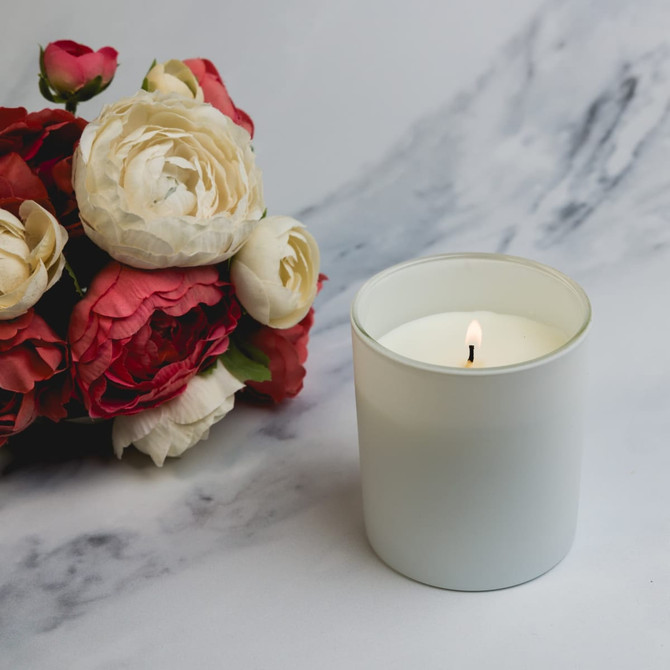 A medium-sized gleam glass candle with a lit wick, surrounded by a bouquet of roses and peonies.