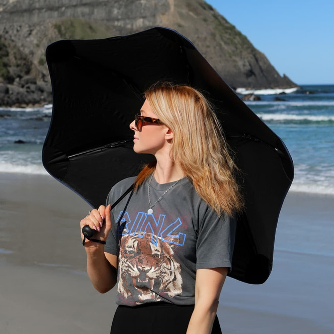 A black BLUNT Metro UV umbrella held by a person on a beach, with waves and cliffs in the background.