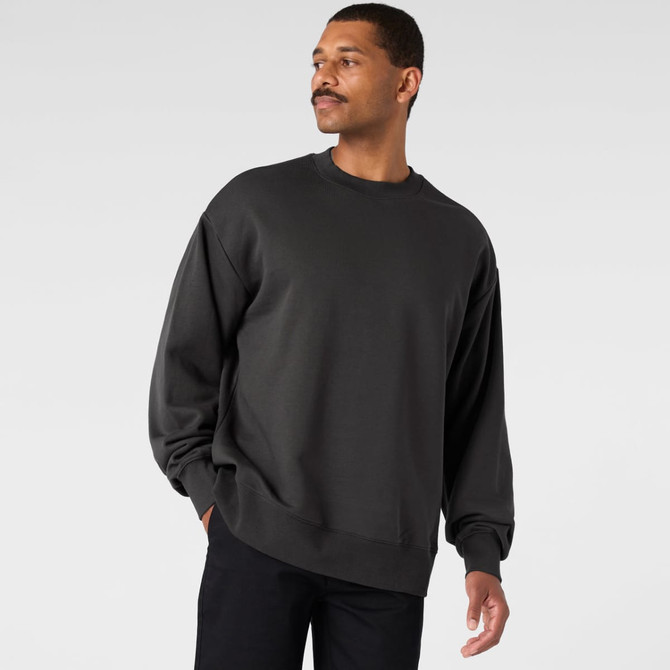 A man wearing a dark grey sweater, standing against a light grey background. The sweater has a relaxed fit and simple design.