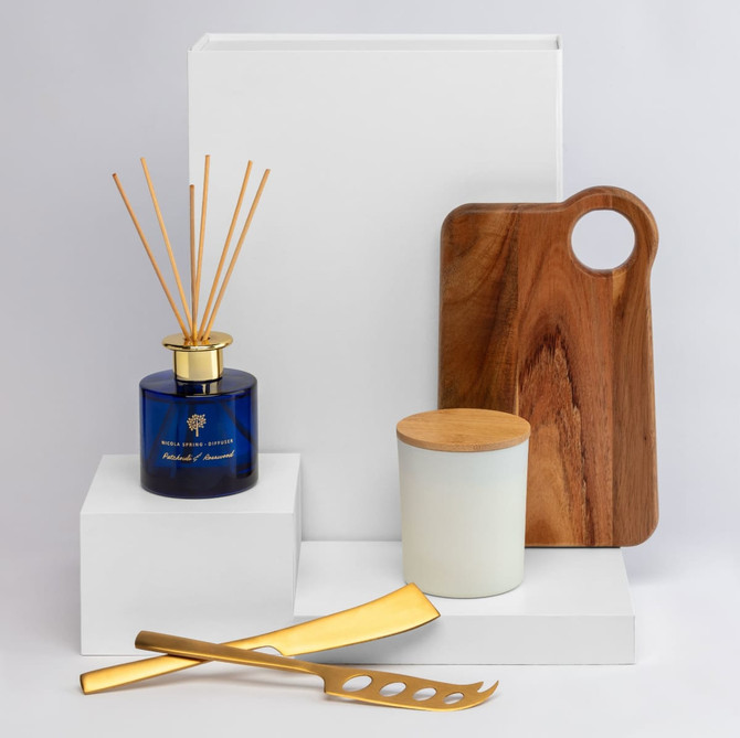 A collection of homeware items including a blue diffuser, wooden board, gold utensils, and a white candle jar on white pedestals.