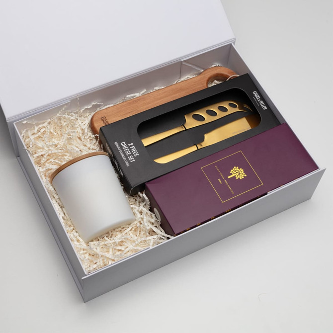 A gift box containing a white candle, wooden spoon, and a black knife set with gold accents, surrounded by crinkle paper.