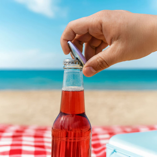 A drink bottle with a silver cap being opened by a hand, set against a beach backdrop and blue sky.