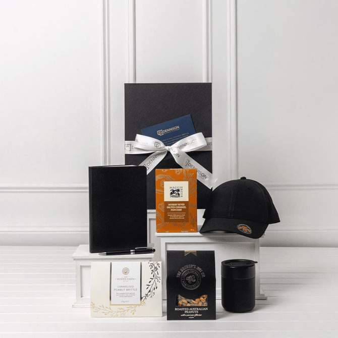 A black gift box with a ribbon and contents including a notebook, pen, coffee cup, cap, and assorted cards.