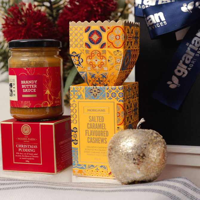 A red jar of brandy butter sauce, a decorative yellow box of salted caramel cashews, and a festive silver apple ornament.