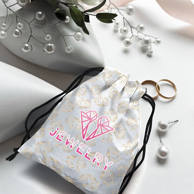 A small white satin gift bag with a drawstring, featuring a pattern and a logo, alongside rings and pearls.