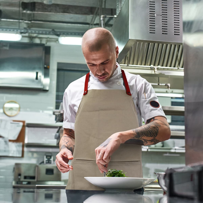 A chef wears a beige bib apron with red straps, preparing a dish in a modern kitchen environment.
