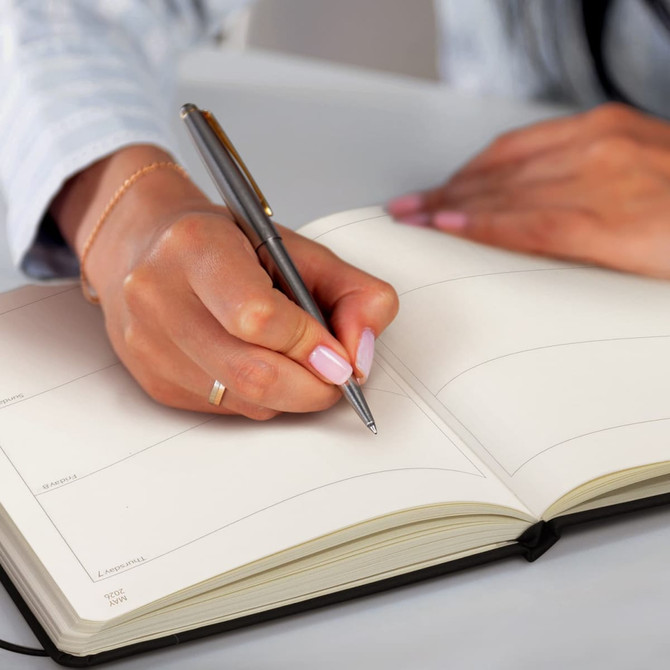 A hand holding a pen writes in a blank weekly diary with cream-coloured pages and a black cover.